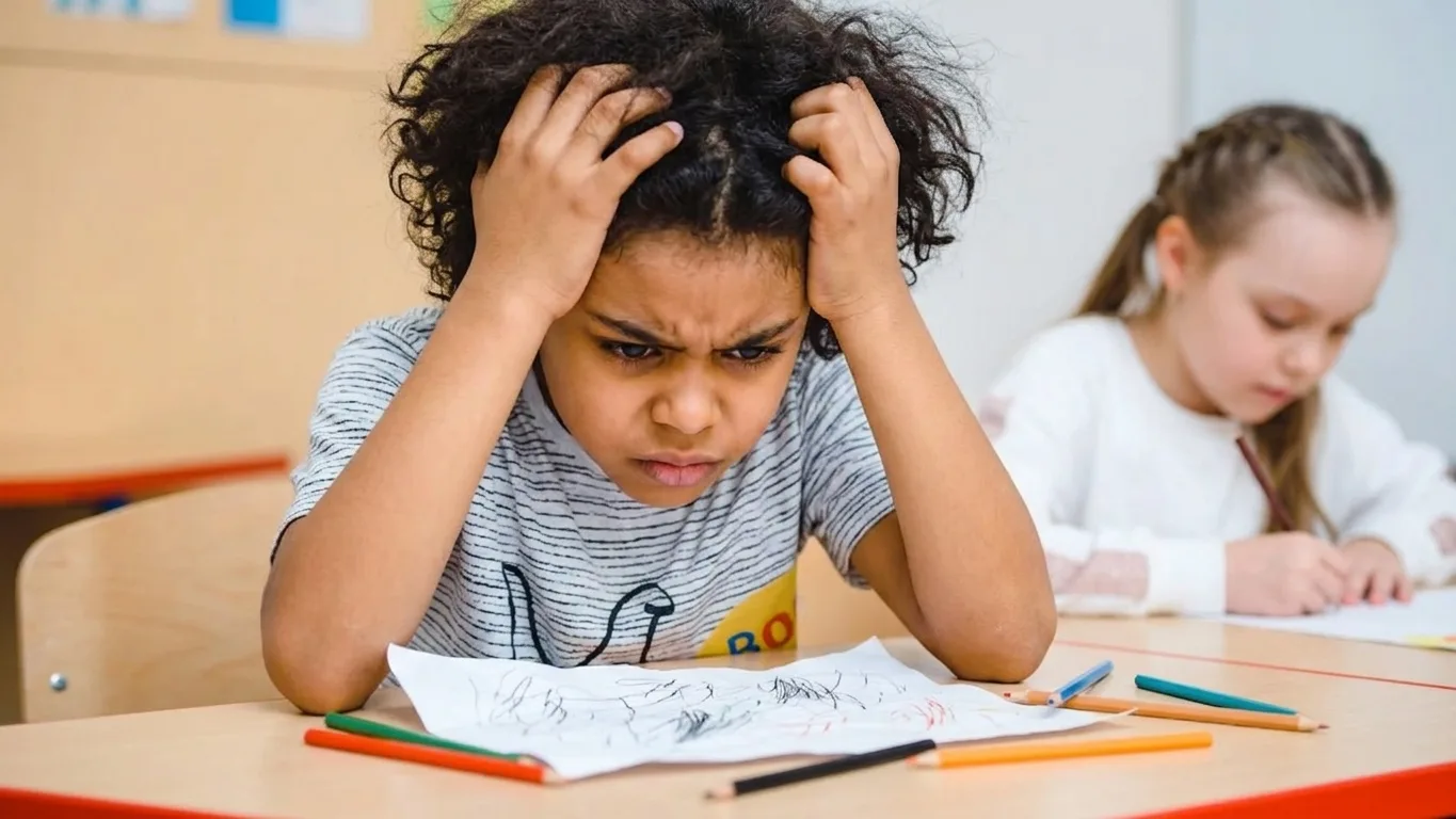 A boy at the kitchen table holding his head over homework while a parent works in the background